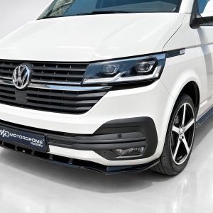 White Volkswagen van front-left view in a showroom, featuring the VW emblem, black grille, and chrome accents with a Motordrome Design plate.