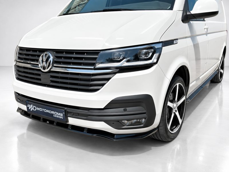 White Volkswagen van front-left view in a showroom, featuring the VW emblem, black grille, and chrome accents with a Motordrome Design plate.
