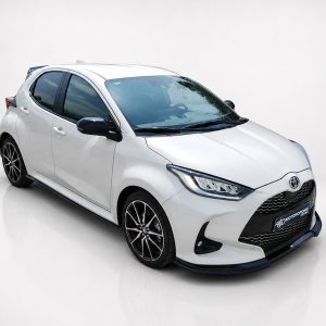 Front-right three-quarter view of a white Toyota hatchback with black mirrors and sporty alloy wheels in a studio setting