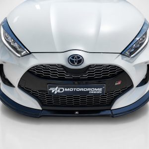 Front view of a white Toyota car showing the grille, emblem, and blue-accented bumper with a Motordrome Design plate at the center bottom of the grill.
