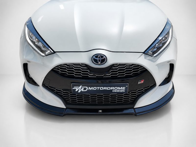 Front view of a white Toyota car showing the grille, emblem, and blue-accented bumper with a Motordrome Design plate at the center bottom of the grill.
