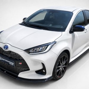 Three-quarter front view of a white Toyota GR hatchback with a black honeycomb grille and blue emblem, accentuated by a sporty black lip and alloy wheels.