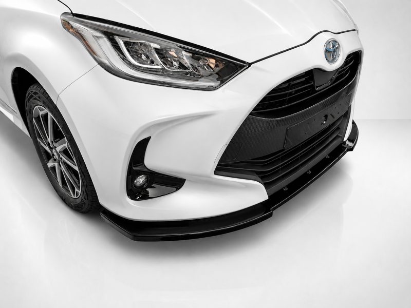 Front-left view of a white Toyota showing the headlight, grille, and front bumper with a carbon-fiber texture detail at the lower grille area.