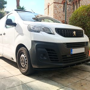 Front three-quarter view of a white delivery van parked on a paved area, with a stone building and greenery in the background, showing the Peugeot-style emblem on the grille.