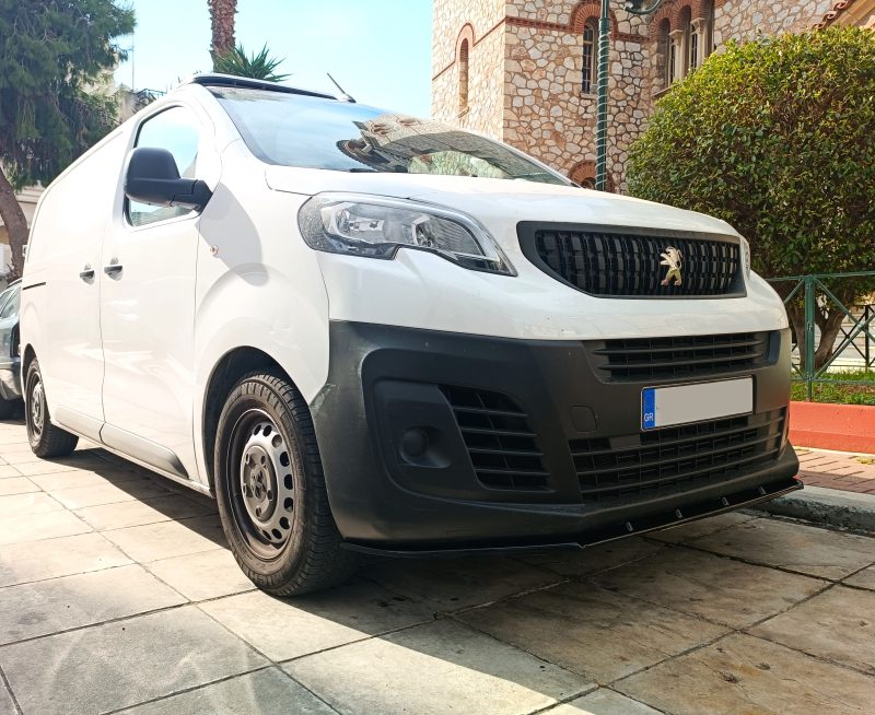 Front three-quarter view of a white delivery van parked on a paved area, with a stone building and greenery in the background, showing the Peugeot-style emblem on the grille.