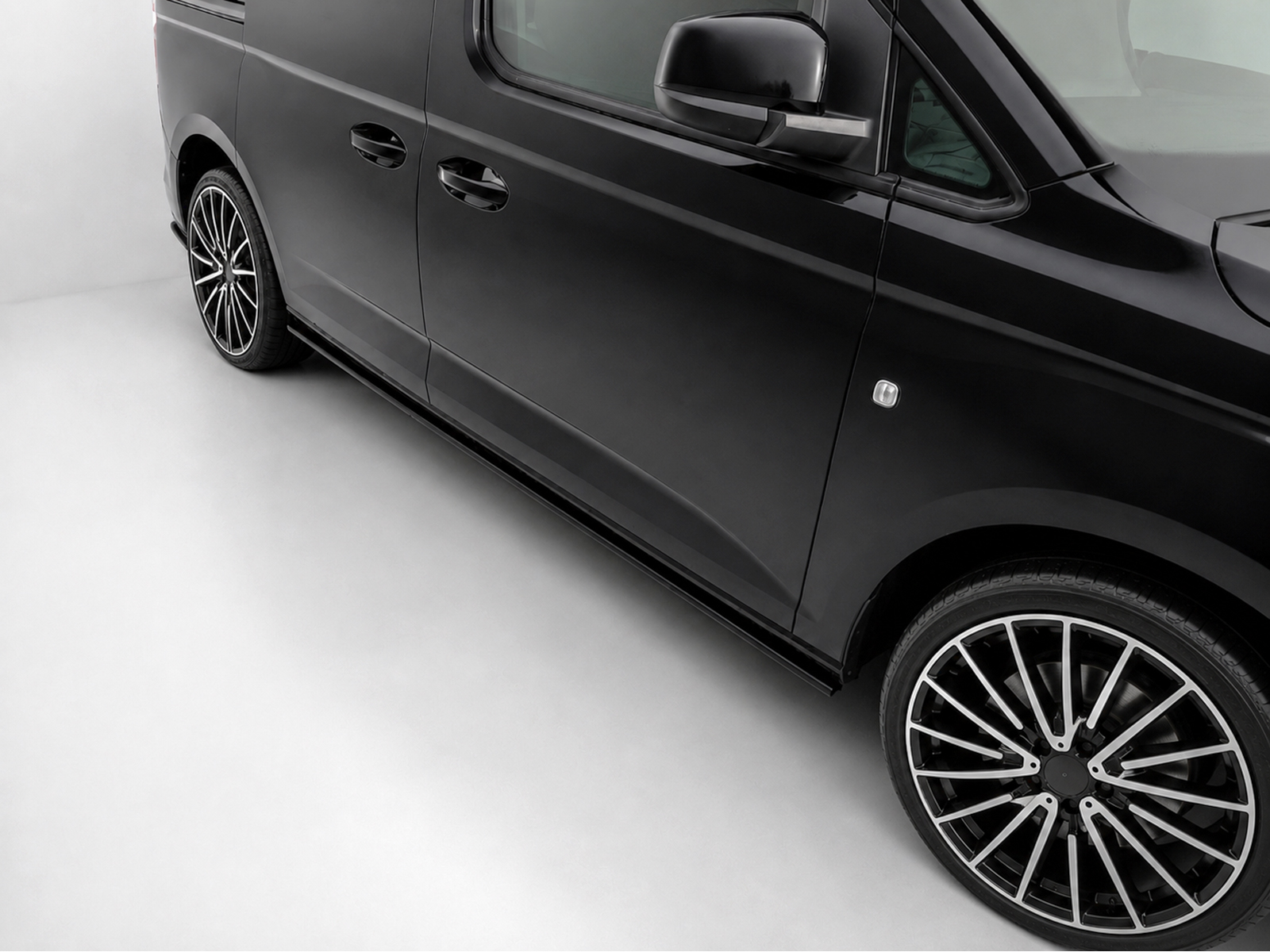 K172-005_2jpg - OPSHOLDERS Close-up of a glossy black van's side with chrome door handles and large multi-spoke alloy wheel on a light-gray floor
