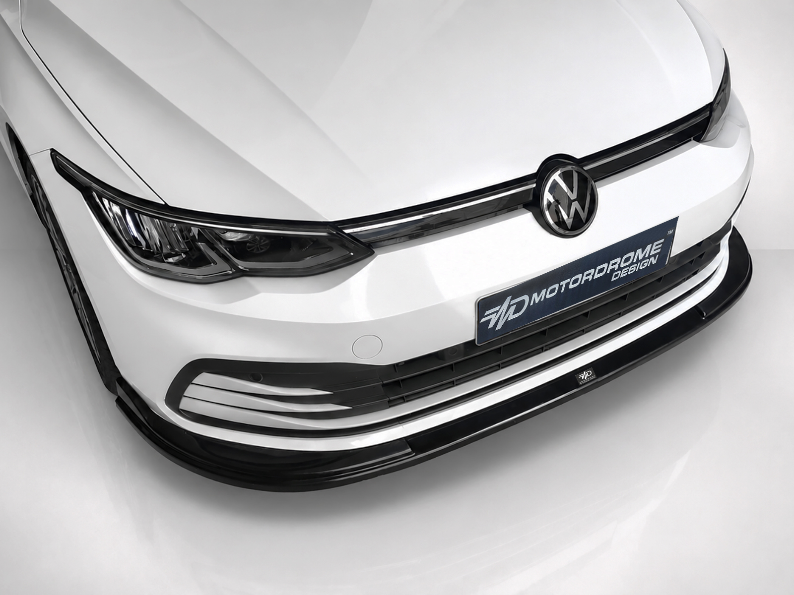 K174-001_1jpg - OPSHOLDERS Close-up of the front end of a white Volkswagen car, showing the emblem, black grille, and a blue license plate reading MOTORODROME DESIGN.