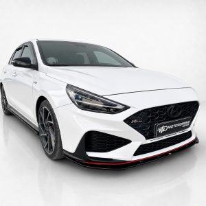 White Hyundai-style sedan photographed from a low front-left angle, showing a black honeycomb grille and red-accent front lip in a studio setting.