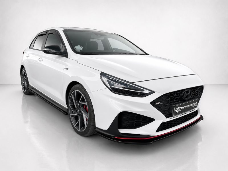 Front three-quarter view of a white Hyundai sporty hatchback with black mesh grille, large dark alloy wheels, and red brake calipers in a studio setting