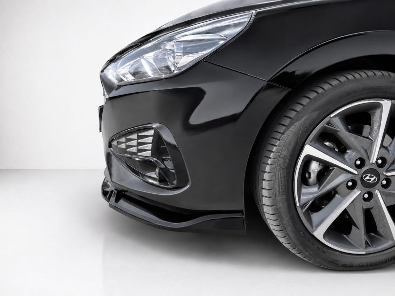 Close-up of a black car's front left corner showing the headlight, fog light area, and alloy wheel in a studio setting.