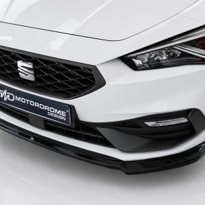 Close-up of the front of a white car, showing a black honeycomb grille, emblem, and sharp headlight with a Motordrome Design plate below the grille.
