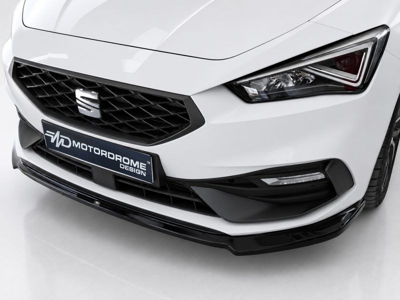 Close-up of the front of a white car, showing a black honeycomb grille, emblem, and sharp headlight with a Motordrome Design plate below the grille.