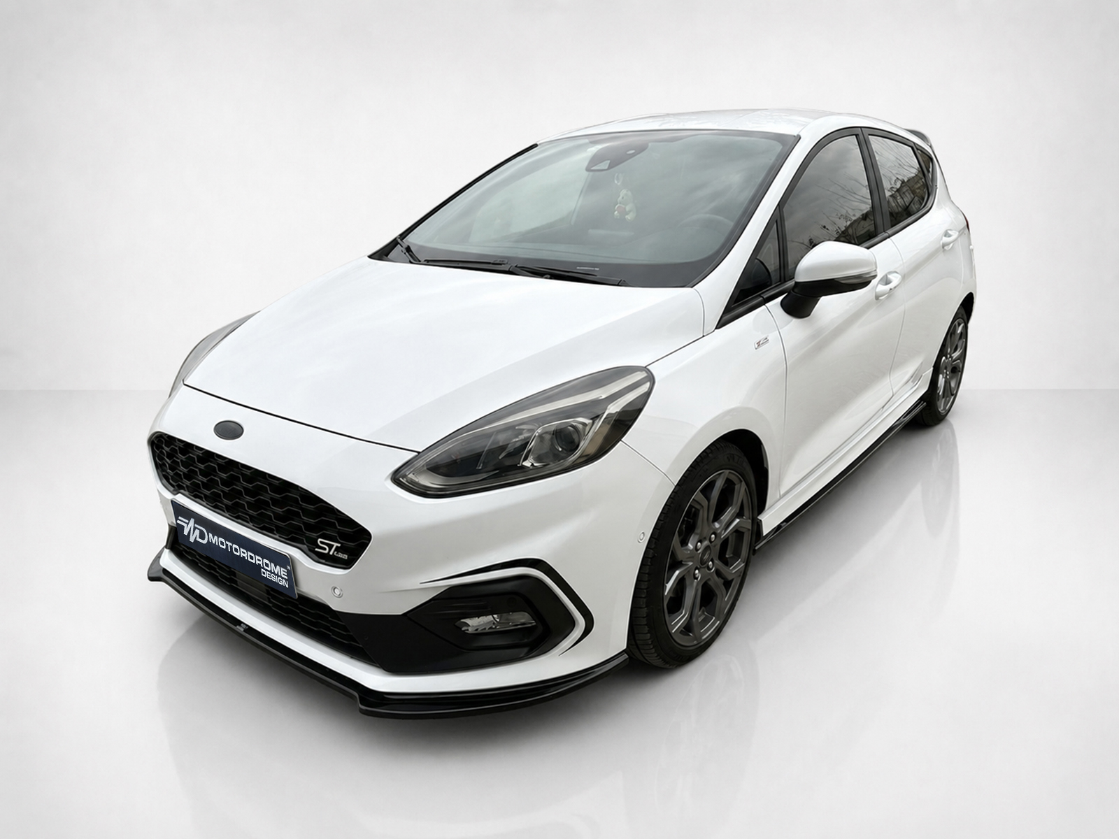 K182-001_1jpg - OPSHOLDERS Angled front-left view of a white sporty hatchback car in a bright studio showroom, showing sleek design and dark alloy wheels.