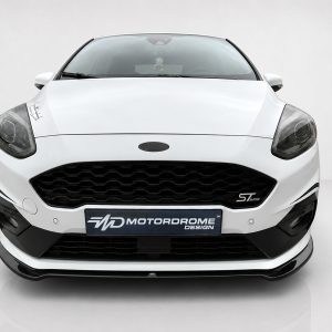 K182-001_2jpg - OPSHOLDERS Front view of a white Ford car in a studio setting, showing honeycomb grille, headlights, and a Motordrome Design license plate.