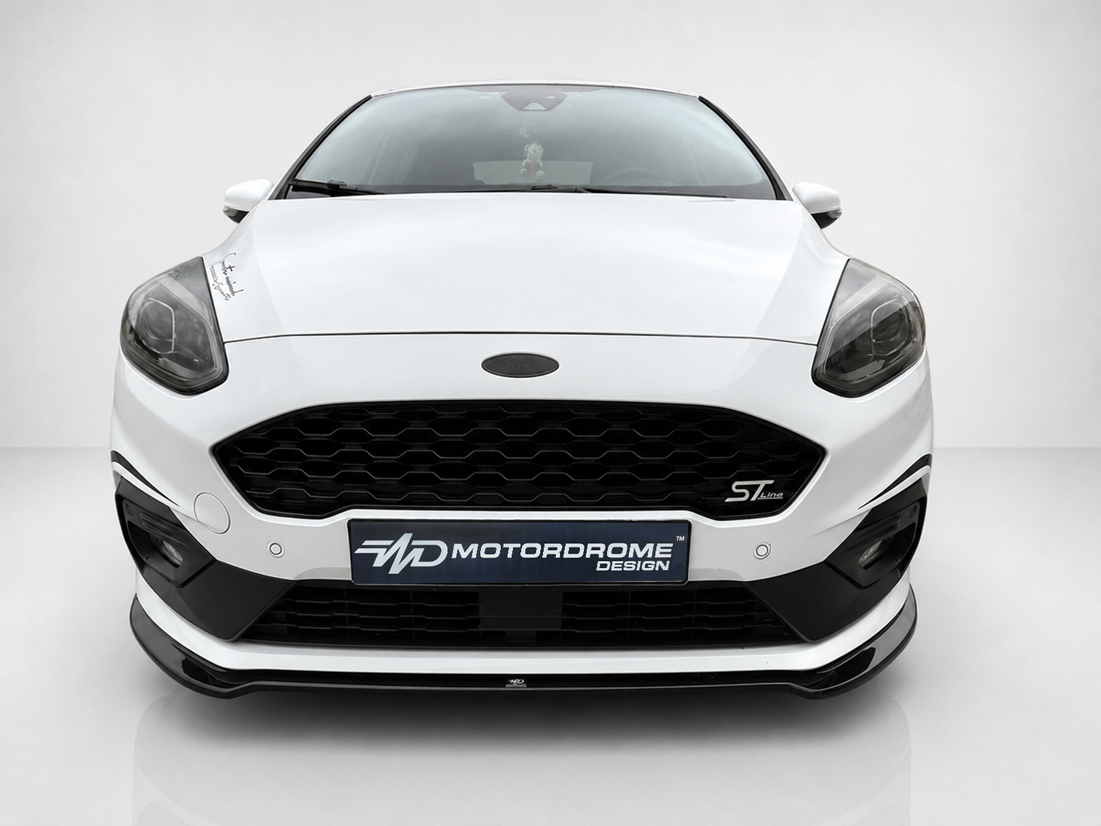 K182-001_2jpg - OPSHOLDERS Front view of a white Ford car in a studio setting, showing honeycomb grille, headlights, and a Motordrome Design license plate.