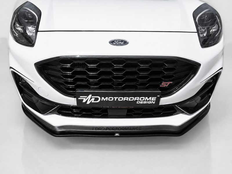 Front view of a white Ford car showing a large black honeycomb grille and Ford badge with a Motordrome Design plate above the bumper edge.