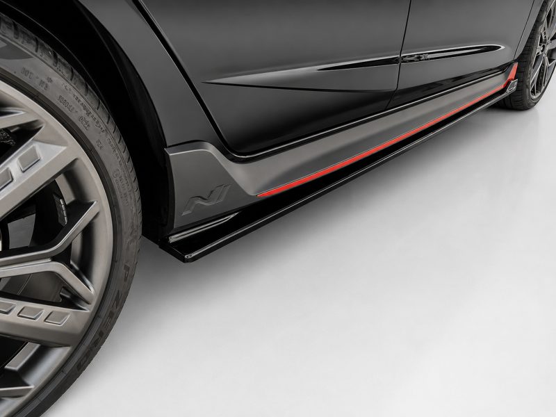 Close-up of a black sports car's lower side panel with a red accent stripe and a large alloy wheel visible on the left.