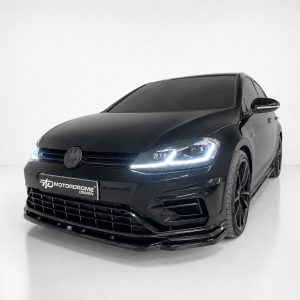 Black VW sedan angled front view in a clean studio with illuminated headlights and a glossy grille; Motordrome Design plate visible