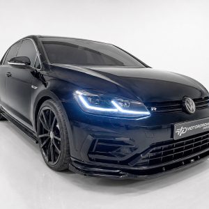 Front three-quarter view of a dark blue Volkswagen with illuminated LED headlights in a white studio, showing large black alloy wheels and Sporty bumper details (VW-R badge visible).