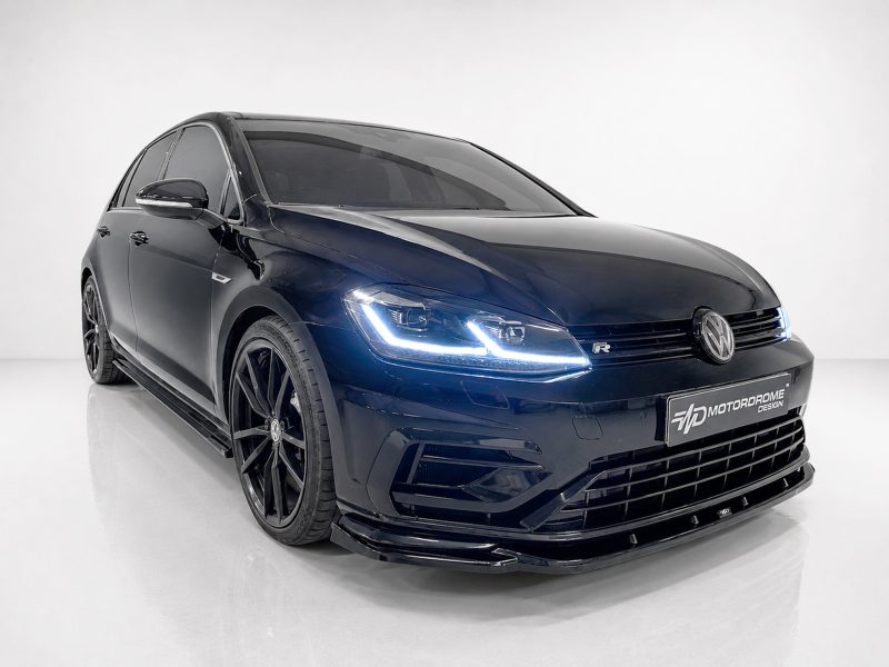 Front three-quarter view of a dark blue Volkswagen with illuminated LED headlights in a white studio, showing large black alloy wheels and Sporty bumper details (VW-R badge visible).