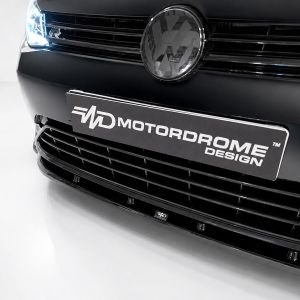 Close-up of a black car front showing a round chrome emblem and a black license plate reading MOTORDOME DESIGN.