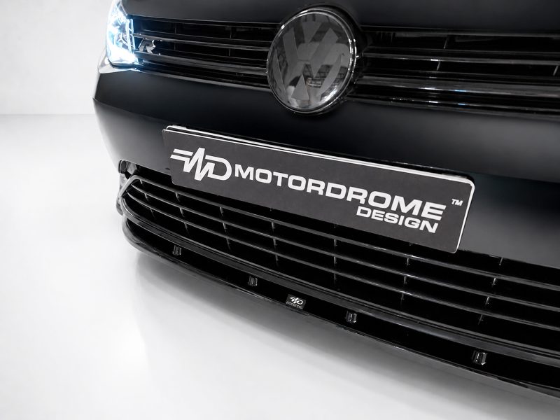 Close-up of a black car front showing a round chrome emblem and a black license plate reading MOTORDOME DESIGN.
