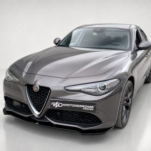 Front three-quarter view of a metallic gray Alfa Romeo sports sedan with a black honeycomb grille and a Motordrome Design plate in a studio setting