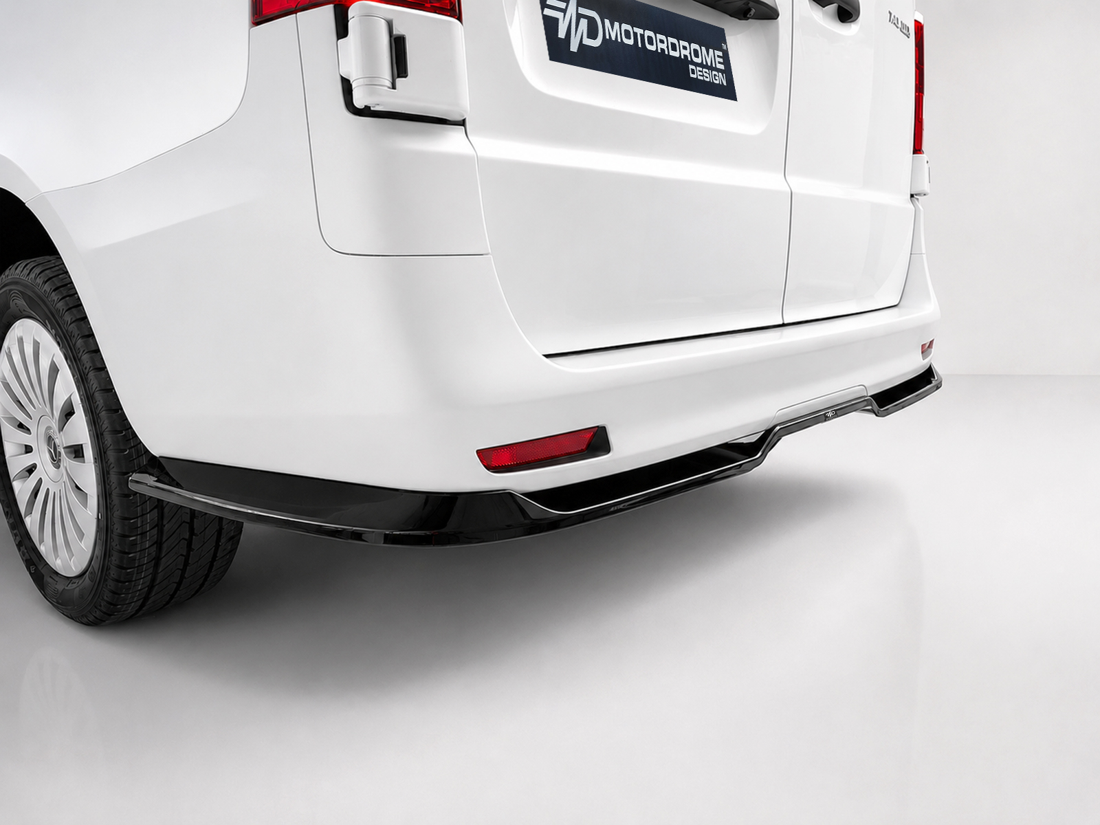 K190-004_1-1jpg - OPSHOLDERS Rear view of a white van with wheel, bumper, and a Motordrome Design badge on the tailgate edge