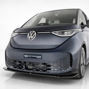 Front-left angled view of a dark blue Volkswagen minivan concept with prominent VW emblem and honeycomb lower grille; showroom backdrop shows 'Motordrome Design' plate.