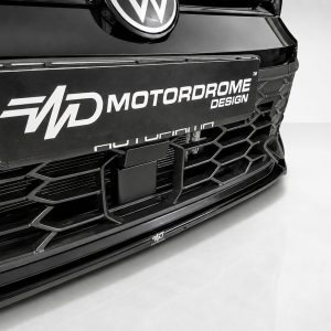 Close-up of a sleek car front with a large rectangular plate reading MOTORODROME DESIGN and a honeycomb grille nearby, in black-and-white.