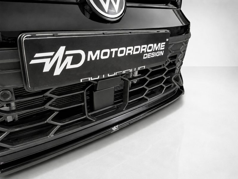 Close-up of a sleek car front with a large rectangular plate reading MOTORODROME DESIGN and a honeycomb grille nearby, in black-and-white.