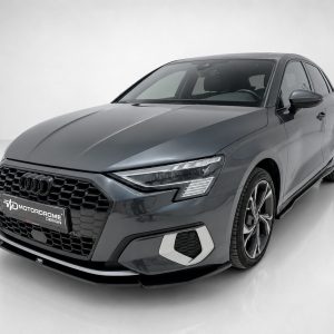 Front three-quarter view of a gray Audi sedan in a studio setting, highlighting the hexagonal grille and LED headlights.