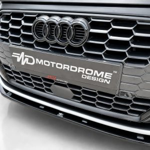 Close-up of a car front grille featuring the Audi rings emblem and a MOTORDROME DESIGN license plate panel.