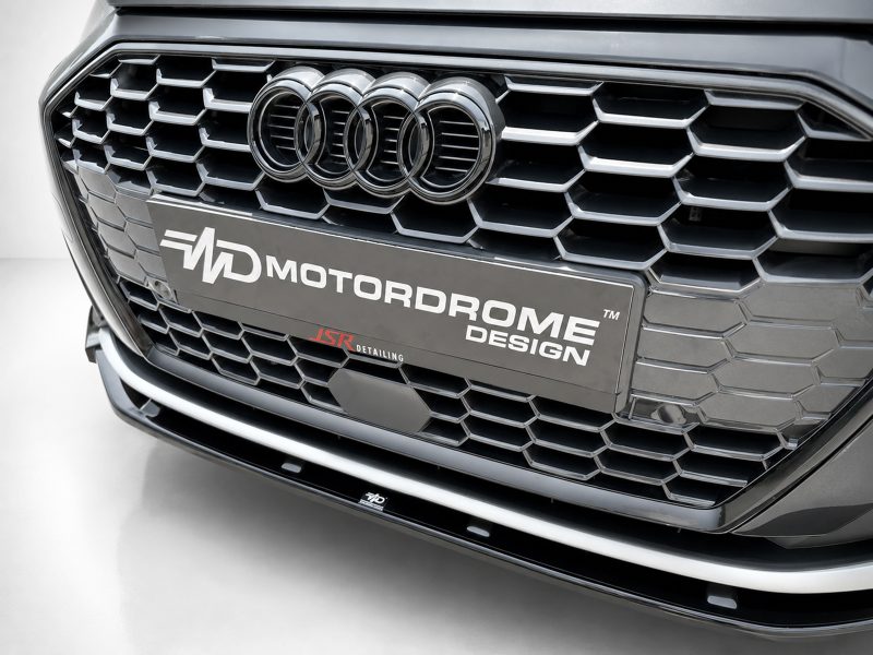 Close-up of a car front grille featuring the Audi rings emblem and a MOTORDROME DESIGN license plate panel.
