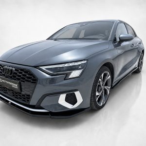 Front three-quarter view of a gray-blue Audi sedan in a studio setting, showcasing a honeycomb grille and sharp LED headlights.