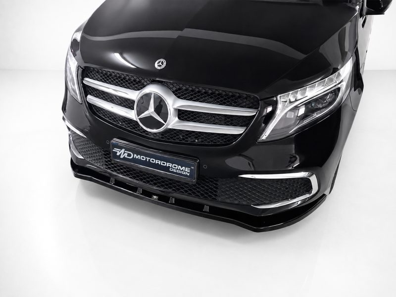 Front view of a black Mercedes-Benz with prominent chrome grille and emblem in a showroom setting, low angle perspective, license plate visible reading MOTORDROME DESIGN