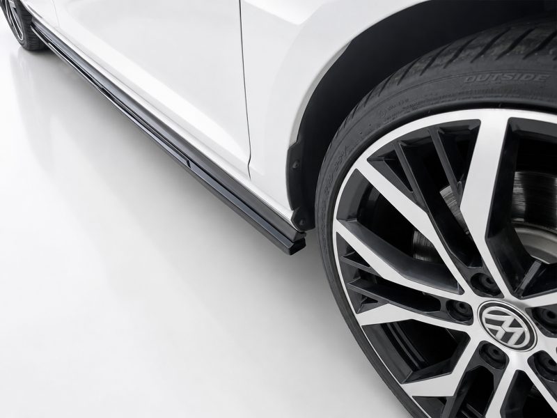 Close-up of a white car's side with a chrome rocker trim and a large black-and-silver alloy wheel visible at the front-right edge, tire labeled outside edge.