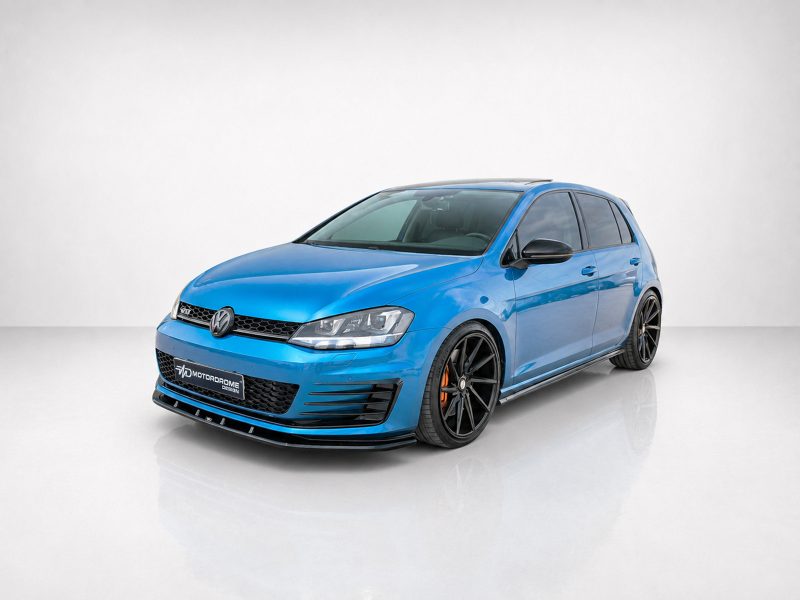 Front three-quarter view of a bright blue modified VW hatchback with black alloy wheels in a studio setting.