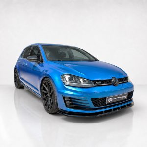 Blue Volkswagen hatchback viewed from the front-right in a studio setting, showcasing a black front lip and large black alloy wheels (aftermarket styling).