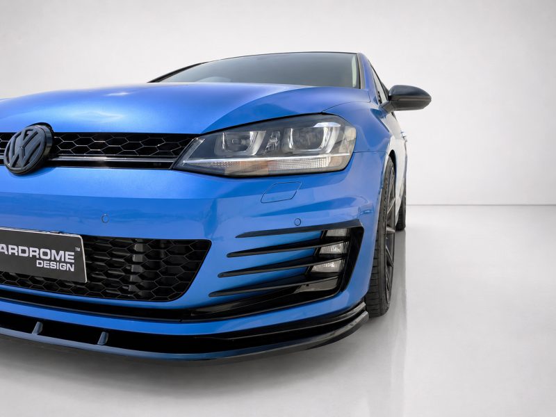 Front-left view of a bright blue Volkswagen with headlight, grille, and lower diffuser visible in a studio setting, license plate partially shown in the corner