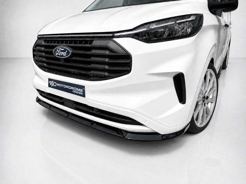 Front-left angle of a white Ford van with a large black grille and badge, displaying a 'MOTORODOME DESIGN' plate at the bumper edge, showroom backdrop