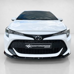Front view of a white Toyota sedan with a black honeycomb grille and prominent logo on the hood emblem, metallic badge visible as 'Motordrome Design' plate
