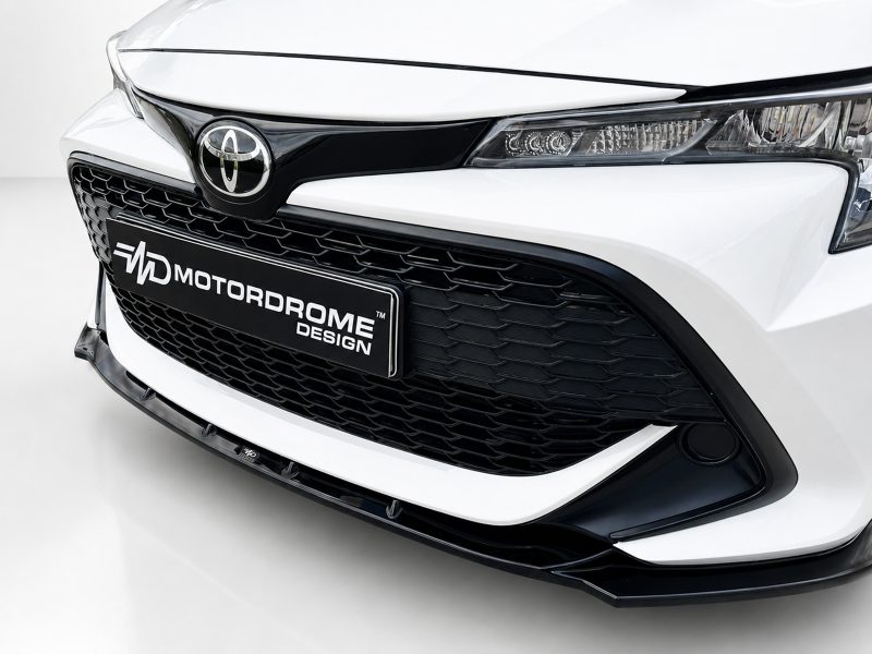 Close-up of a white car's front end with a black honeycomb grille and a license plate reading MOTORDROME DESIGN.