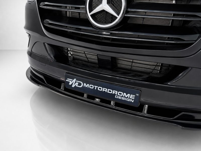 Close-up of the front of a glossy black car showing the grille, Mercedes emblem, and a MotorDome Design plate nearby.