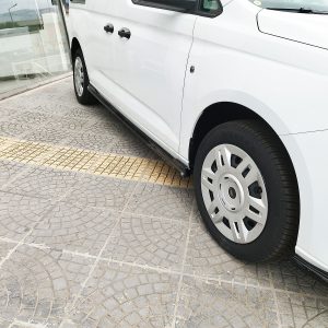 White van parked beside a glass storefront, showing the side step and front wheel on a patterned pavement surface.