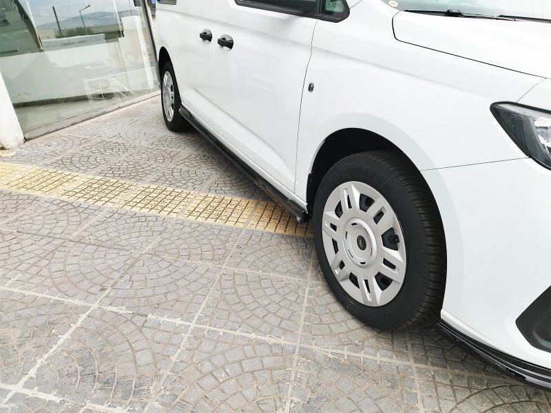 White van parked beside a glass storefront, showing the side step and front wheel on a patterned pavement surface.