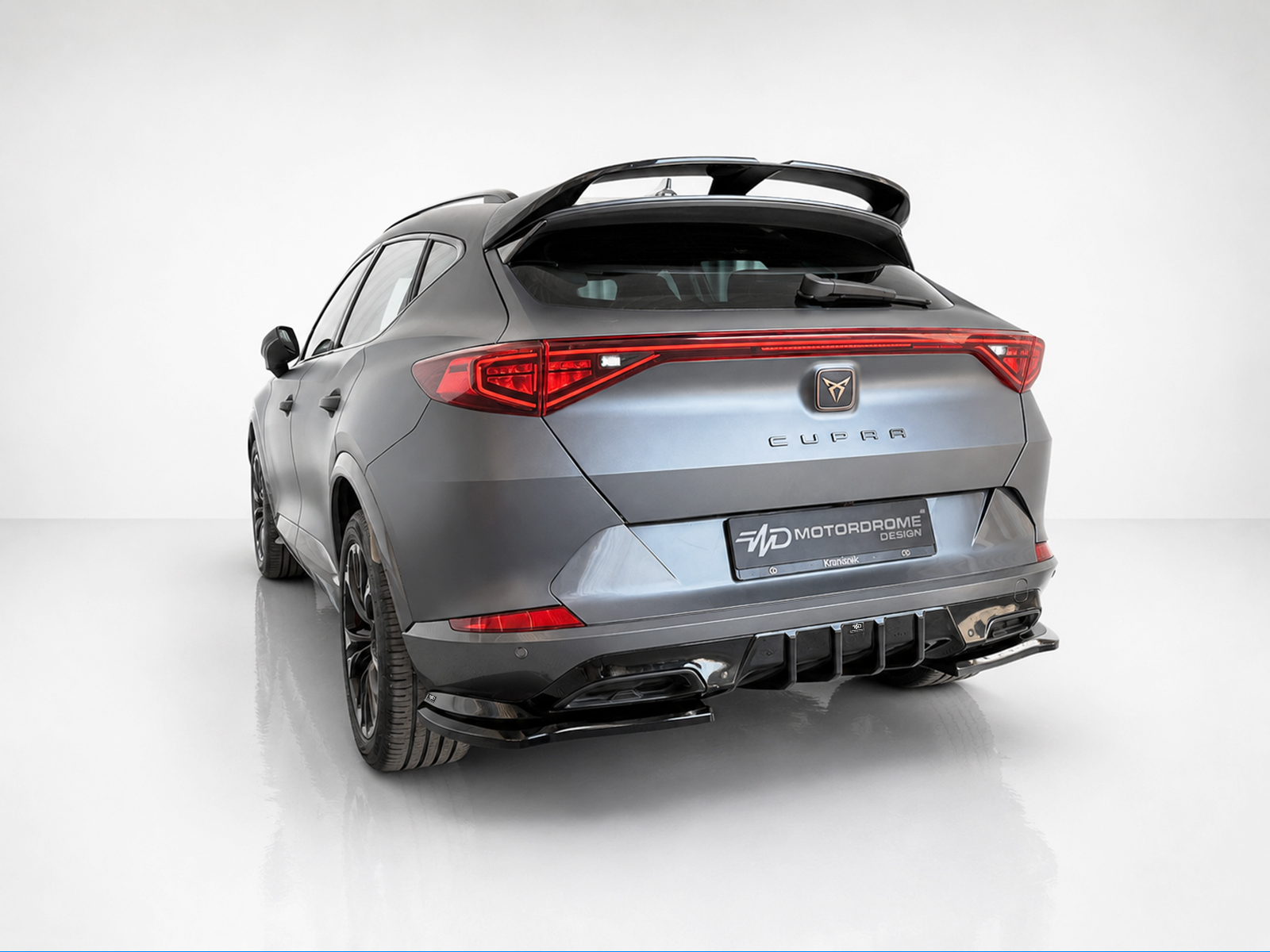 K208-003_1jpg - OPSHOLDERS Rear three-quarter view of a metallic gray CUPRA hatchback/SUV with a large rear spoiler in a white studio setting, showing red taillights and badge row.