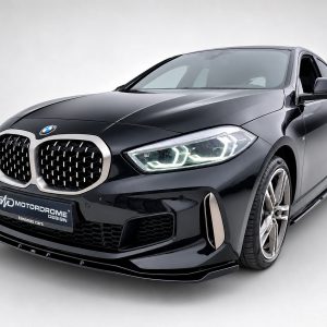 Front three-quarter view of a glossy black BMW sedan with a prominent chrome kidney grille and illuminated headlights, indoors on a light background.