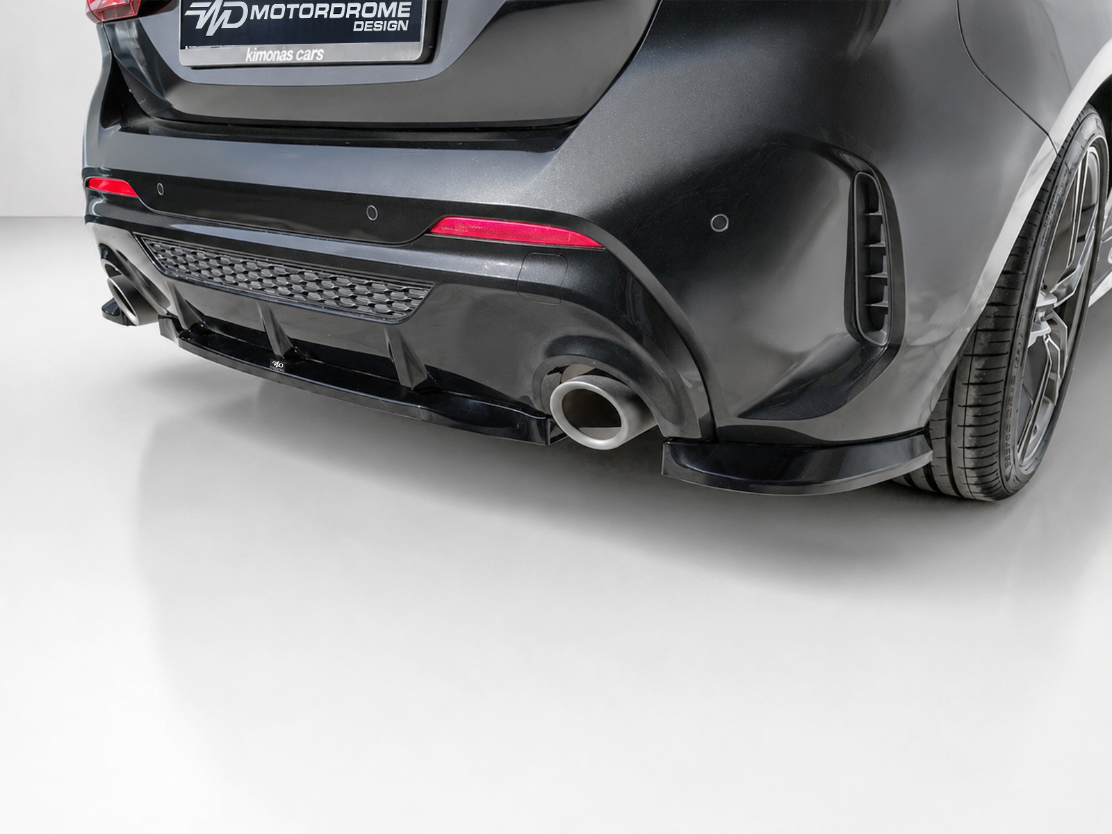 K210-003_2jpg - OPSHOLDERS Rear view of a dark sports car showing a large exhaust, diffuser, and low bumper with red taillights and a wide wheel visible on the right edge.