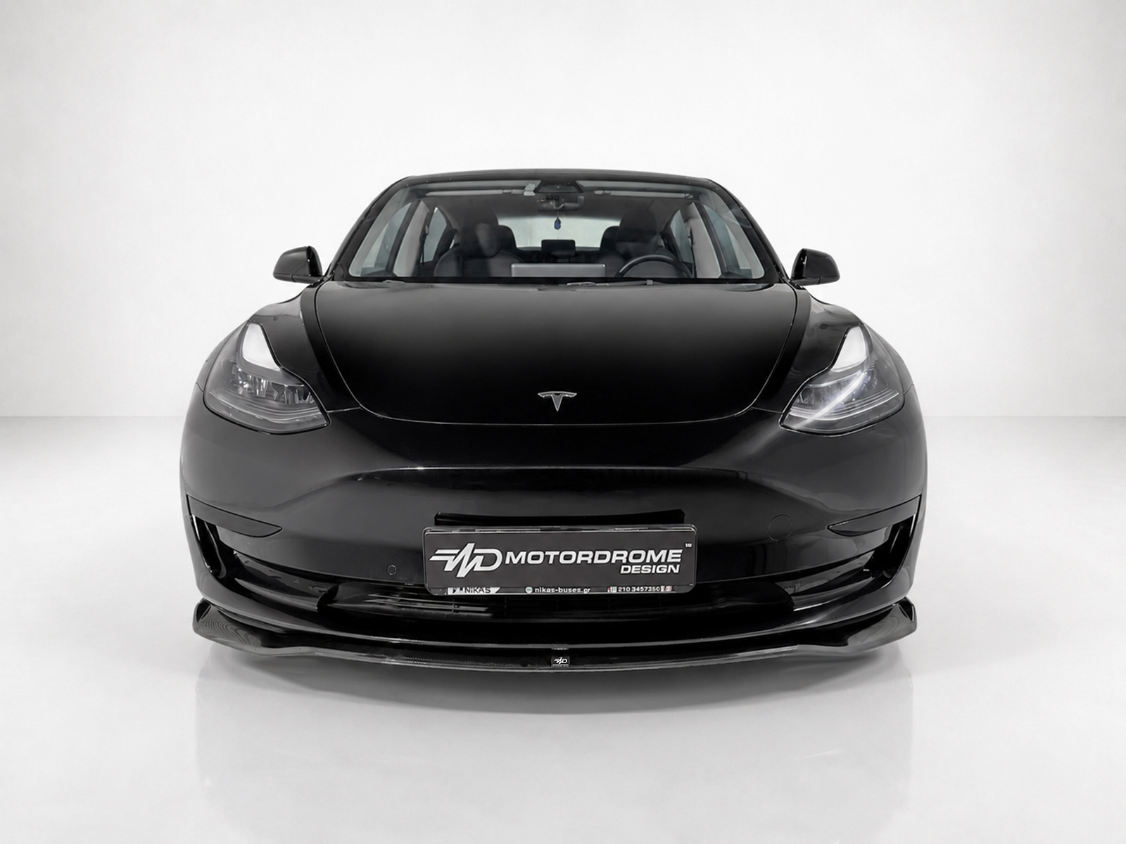 K214-001_1jpg - OPSHOLDERS Front view of a black Tesla car in a white studio, with a Motorodrome Design plate on the bumper
